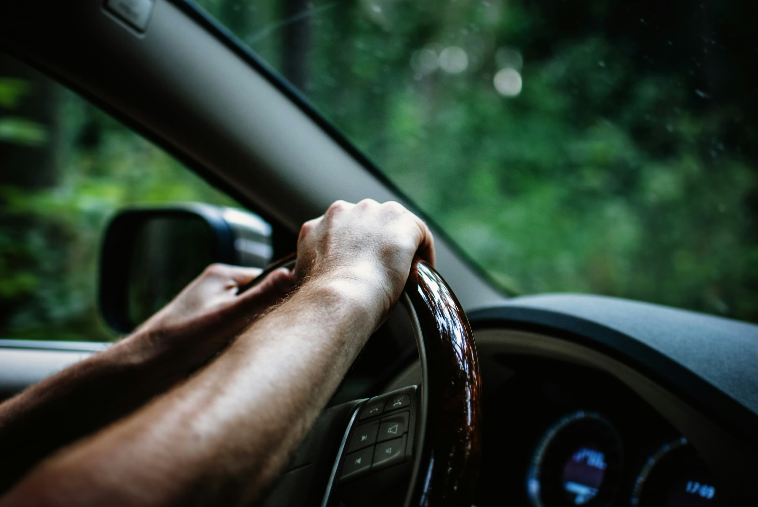 A driver's hand on the steering wheel with soft green woodland visible through the window